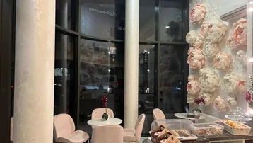 Innenbereich eines Cafés mit großen Fenstern, rosa Stühlen und einer Theke mit Gebäckauslage.
