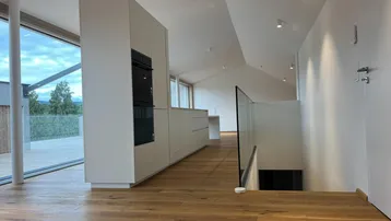 Moderne offene Küche mit integriertem Backofen, Holzboden und großen Fenstern mit Blick ins Grüne.