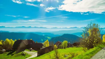 Weitläufige Landschaft mit traditionellen Häusern auf einem Hügel und majestätischem Bergblick unter blauem Himmel.
