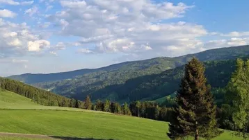 Panoramablick auf grüne Hügel und bewaldete Berge unter blauem Himmel mit weißen Wolken.