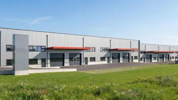 Modernes Industriegebäude mit mehreren Eingängen und großen Fenstern, umgeben von grünen Wiesen unter blauem Himmel.