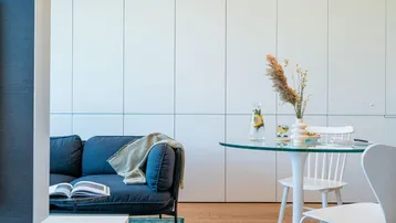 Moderner Wohnbereich mit blauem Sofa, Glastisch und integrierten weißen Schränken auf hellem Holzfußboden.