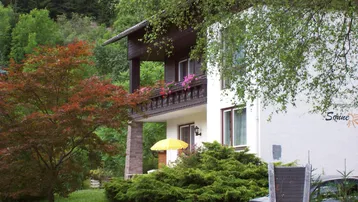 Traditionelles Haus mit Holzbalkon und grüner Umgebung, umgeben von Bäumen und Natur.