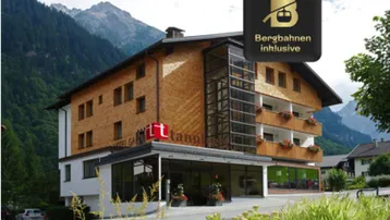Modernes Gebäude mit Holzfassade, Glasaufzug und Balkonen, umgeben von einer beeindruckenden Berglandschaft.