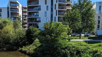 Moderne Wohngebäude am Flussufer mit üppiger Vegetation unter blauem Himmel.