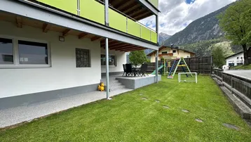 Außenansicht des Gebäudes mit großem Garten, Spielplatz und gemütlicher Terrasse. Im Hintergrund ist ein beeindruckender Bergblick zu sehen.