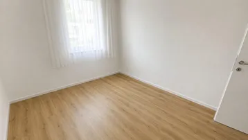 Helles, leeres Zimmer mit Laminatboden und einem Fenster, das viel Tageslicht hereinlässt.
