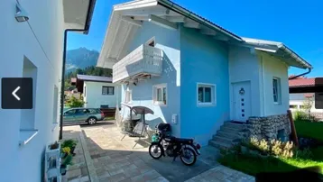 Blau gestrichenes Einfamilienhaus mit weißem Balkon und gepflastertem Eingangsbereich.