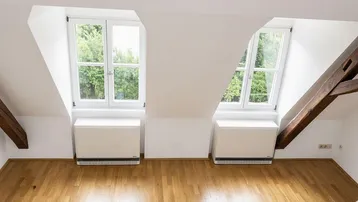 Helles Dachzimmer mit Holzboden, schrägen Wänden und zwei Fenstern mit Blick ins Grüne.