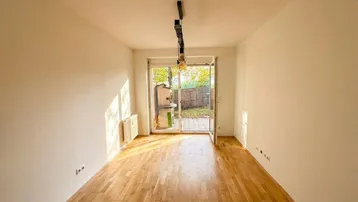 Helles, leeres Wohnzimmer mit Holzboden und direktem Zugang zu einem kleinen Gartenbereich.