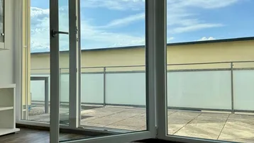 Heller Innenraum mit großen Fenstern und direktem Zugang zu einer geräumigen Terrasse unter blauem Himmel.