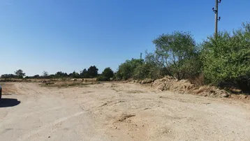 Eine weite, unbefestigte Fläche unter blauem Himmel, umgeben von spärlicher Vegetation und Strommasten.