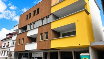 Moderne Wohnanlage mit Holzfassade, gelben Akzenten und Balkonen unter blauem Himmel.