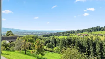 Weitläufiger Blick über grüne Hügel, Wälder und eine Stadt am See unter blauem Himmel.