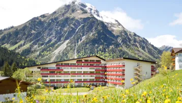 Großes Gebäude mit roten Balkonen und schneebedecktem Berg im Hintergrund, umgeben von grünen Wiesen.
