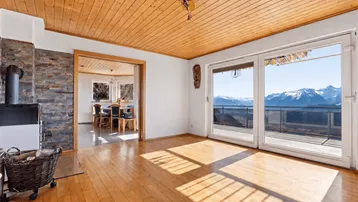 Gemütliches Wohnzimmer mit Holzboden, Kaminofen und Zugang zu einem Balkon mit Bergblick.