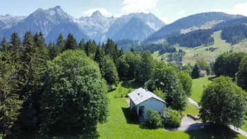 Luftaufnahme eines Hauses in einer grünen Berglandschaft mit vielen Bäumen und klarem Himmel.