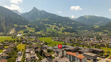 Luftaufnahme der Ortschaft mit umliegenden Bergen und markiertem Gebäude unter blauem Himmel.