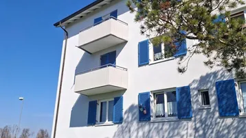 Weißes Mehrfamilienhaus mit blauen Fensterläden und Balkonen in ländlicher Umgebung.