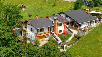 Luftaufnahme eines Hauses mit roter Umrandung, umgeben von grünen Hügeln und Bäumen.