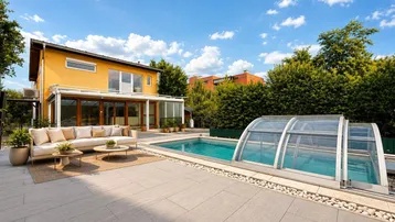 Große geflieste Terrasse mit Pool, bequemen Gartenmöbeln und einem Haus mit gelber Fassade unter blauem Himmel.