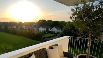 Geräumiger Balkon mit bequemen Sitzmöbeln, Olivenbaum und weitem Blick über grüne Felder bei Sonnenuntergang.