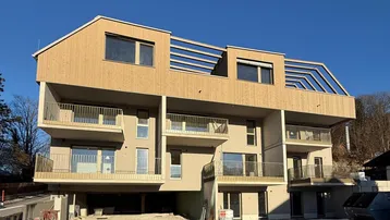 Modernes Mehrfamilienhaus mit Holzfassade und mehreren Balkonen, umgeben von einer Baustelle.