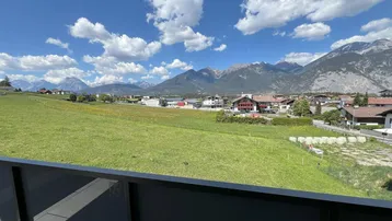 Panoramablick von einem Balkon über grüne Felder auf ein Dorf und die umliegende Berglandschaft.