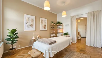 Heller Behandlungsraum mit Massageliege, Holzregal und Fischgrätparkett, ideal für Wellness oder Therapie.