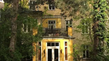 Gelbe Fassade eines Hauses mit Balkon und üppigem Gartenbereich, umgeben von hohen Bäumen.