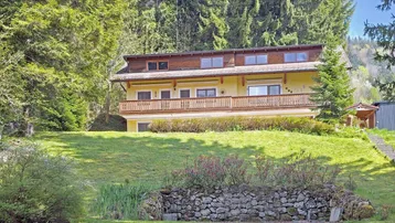 Hausfassade mit mehreren Balkonen, umgeben von viel Grün und Bäumen auf einem Hanggrundstück.