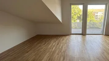 Helles Zimmer mit Dachschräge, Holzboden und Zugang zu einem Balkon mit Blick ins Grüne.