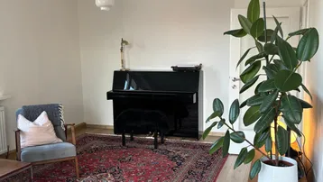 Geräumiges Wohnzimmer mit hellem Parkettboden, einem Teppich und einem Klavier.