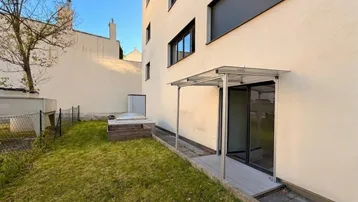 Außenbereich mit Rasenfläche, überdachtem Terrassenzugang und Hochbeet am modernen Gebäude.