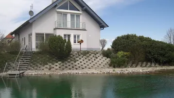 Modernes Haus mit Giebeldach, Balkon und einem natürlichen Teich im Vordergrund, umgeben von Grün.