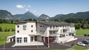 Modernes Gebäude mit Balkonen und Bergblick, umgeben von grünen Wiesen unter blauem Himmel.