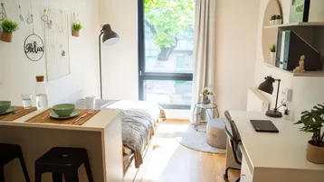 Helles Studio-Apartment mit Schlafbereich, Schreibtisch und Essgelegenheit am Fenster.