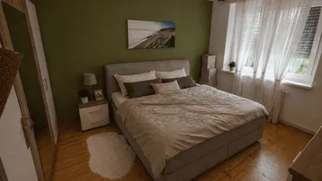 Helles Schlafzimmer mit großem Doppelbett, grüner Akzentwand, Holzfußboden und großem Fenster mit Rollladen.