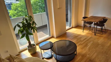Helles Wohn- und Esszimmer mit Holzboden, Zugang zum Balkon und Blick ins Grüne.