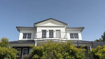 Modernes weißes Haus mit Balkon und dunklem Satteldach unter klarem blauen Himmel, umgeben von grünen Büschen.
