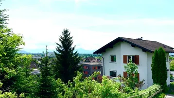 Hausfassade mit grüner Umgebung und Fernblick auf die Landschaft unter blauem Himmel.