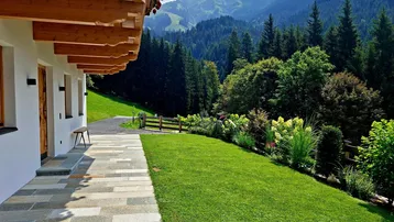 Seitlicher Blick auf das Haus mit gepflasterter Terrasse, Rasenfläche und beeindruckendem Bergblick im Hintergrund.