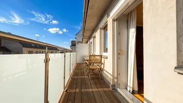 Langer Balkon mit Holzdeck, Glasgeländer und Sitzgelegenheiten unter blauem Himmel.