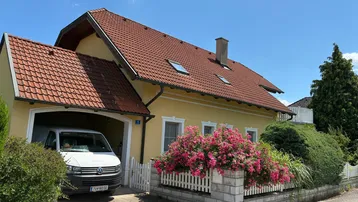 Gelbes Einfamilienhaus mit rotem Ziegeldach, Garage und blühenden Sträuchern im Vorgarten.