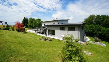 Modernes Wohnhaus mit überdachter Terrasse und weitläufigem Garten in sonniger Hanglage.