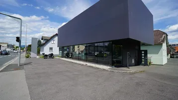 Modernes Geschäftsgebäude mit großer Glasfassade und Parkplätzen an einer Straße unter blauem Himmel.