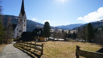 Weitläufige Landschaft mit Kirche, Wiesen und Bergen unter strahlend blauem Himmel.