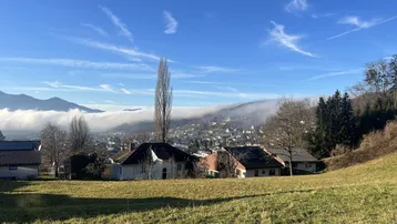 Weitläufiger Blick über eine grüne Wiese auf eine neblige Talsiedlung mit Bergen im Hintergrund unter blauem Himmel.