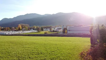 Sonnige Landschaft mit weiten grünen Feldern, Bergen im Hintergrund und vereinzelten Gebäuden.