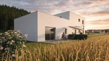 Modernes weißes Haus mit Flachdach und Terrasse, umgeben von Feldern und Wald bei Sonnenuntergang.